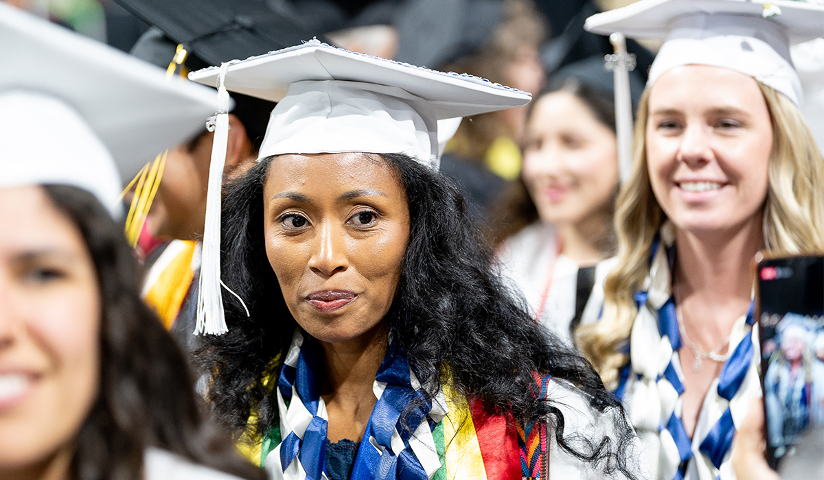 A nursing graduate wearing a white cap and gown sits among other graduates.