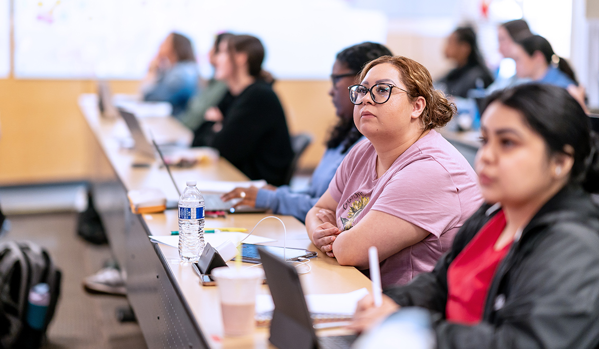 A student sits at a table in a lecture hall and listens to an instructor.